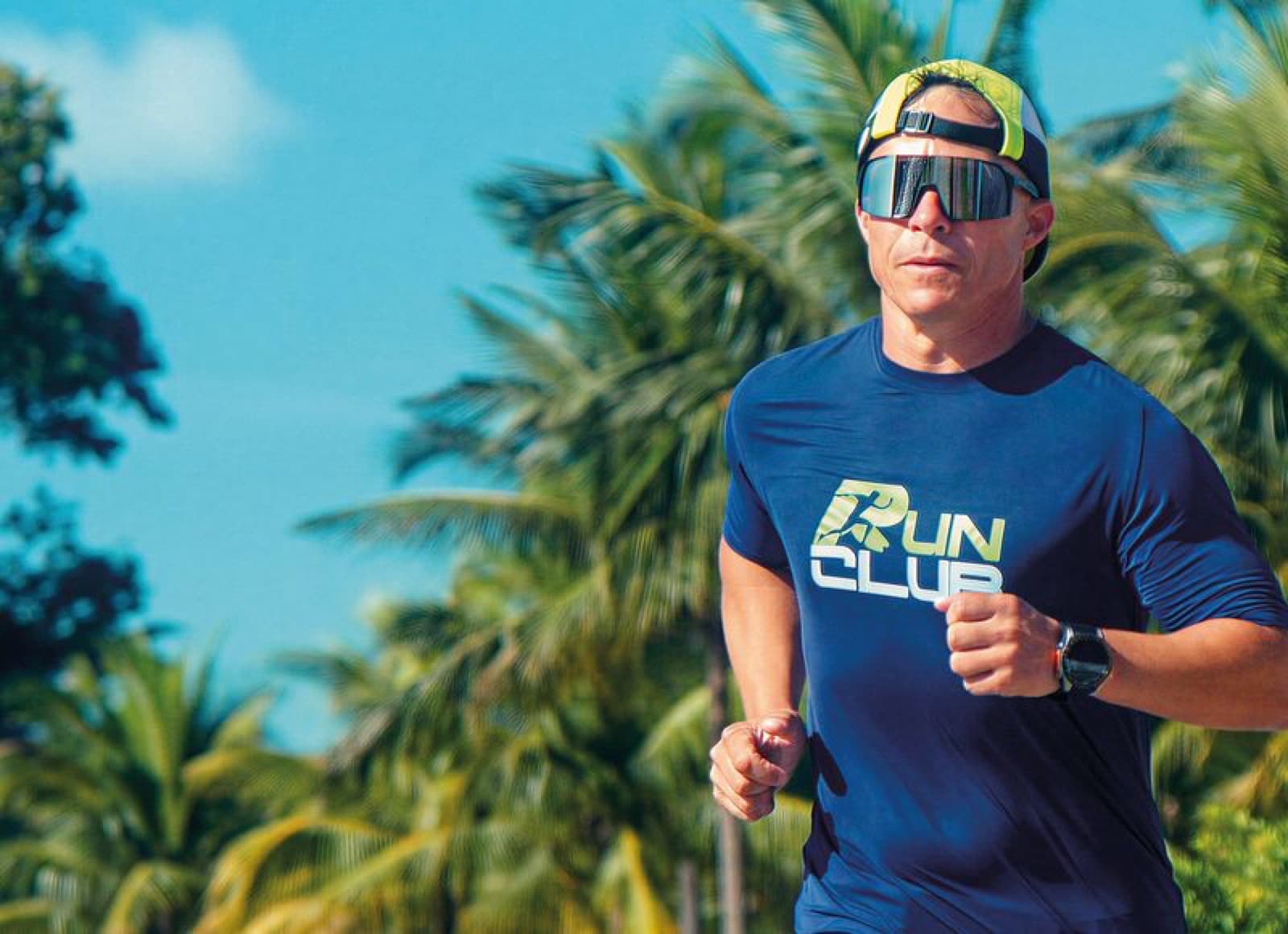 Edinho correndo entre coqueiros num dia de sol, de óculos escuros e camisa azul da RunClub