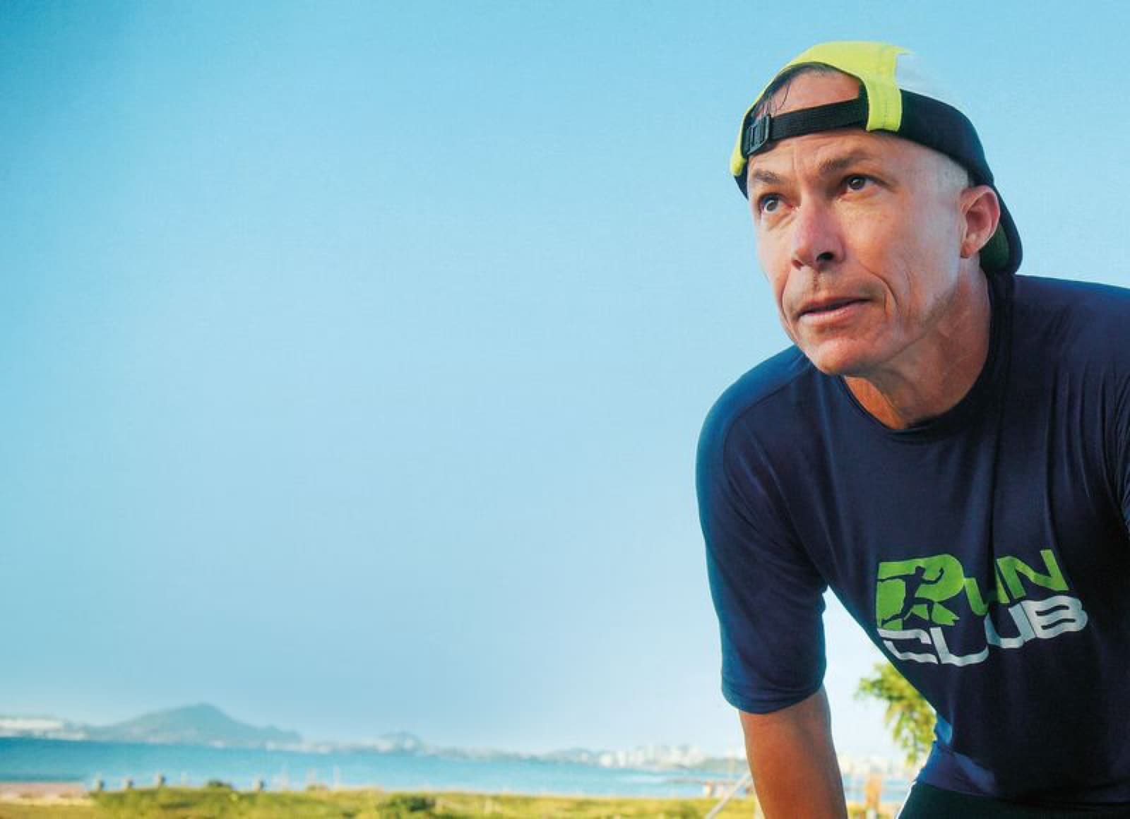 Edinho de boné virado pra trás e camisa da RunClub, inclinado pra frente, olhando pro horizonte com o mar e a cidade ao fundo